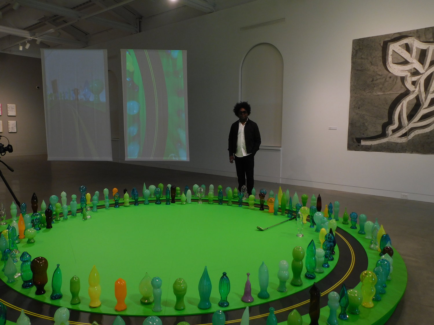 “Landscape and Hierarchies” by Alexandre Arrechea is an installation of a circular putting green inlaid with a track for a golf ball amid a forest of glass-blown sculptures of trees. The piece is flanked by video projections that offer alternative views of the imagined
landscape.