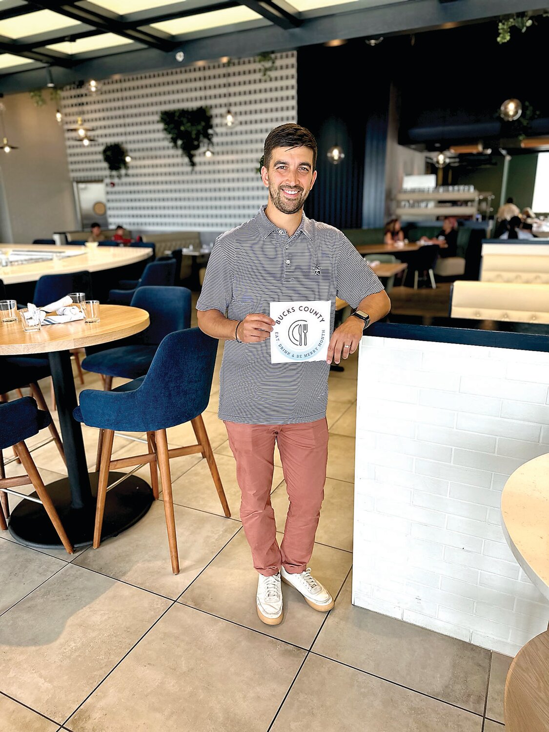 Mike Christou, owner of Ardana Food and Drink, holds up the Bucks County Eat, Drink and Be Merry Month logo. All participating locations will receive a partnership decal to display on the window of their business.