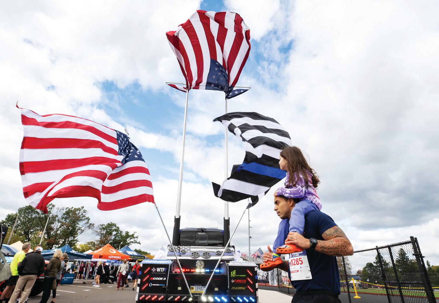 A participant in the 9/11 Heroes Run walks beneath three flags mounted on the back of a truck.