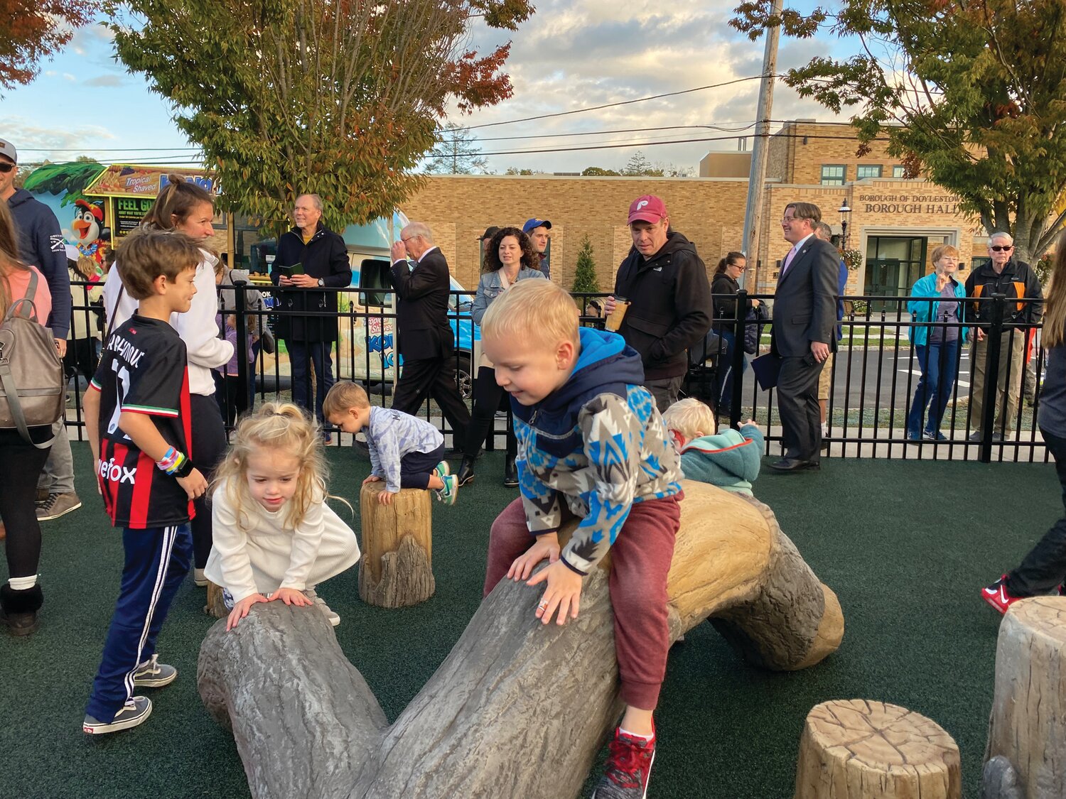 As parents watch from the fence, children climb toward a slide that resembles a huge tree trunk, with faux logs serving as steps.