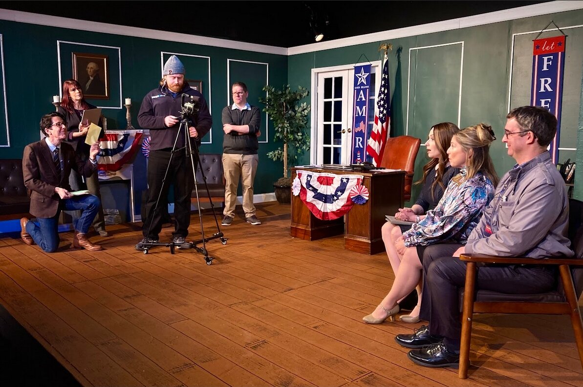 From left are Joseph Torsella, Cathy Liebars, Esh Ryans, John Boccanfuso, Cat Miller, Shelli Pentimall Bookler, and Nigel Rogers in a press conference scene from “The Outsider.”
