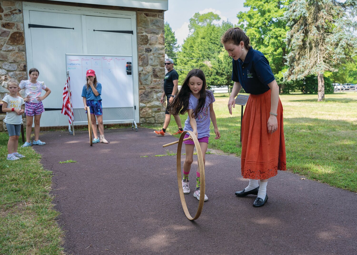 Corrine Kauermann, historical interpreter, educational programming team, helps Polly Parks with the hoop and stick as, from left, Iris Warren, Celeste O’Toole, Tara O’Toole and James Warren look on.