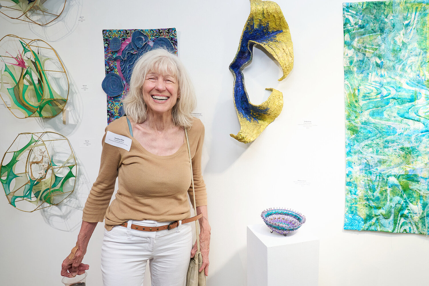 Exhibiting Fiber Artist Sandra Eliot standing among her art and other artists’ work during the exhibit’s opening night.